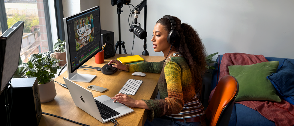 A woman in her home working at her desk with MacBook Pro connected to two external ekraans, wearing headphones and speaking into a microphone