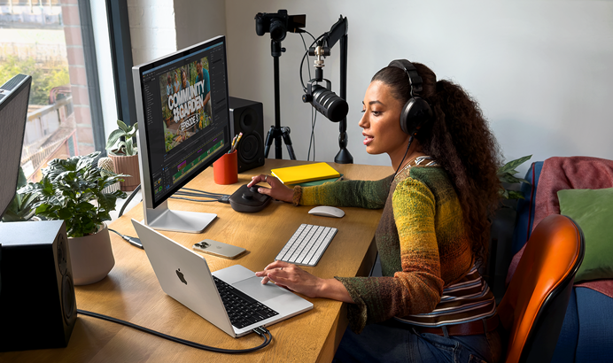 A woman in her home working at her desk with MacBook Pro connected to two external ekraans, wearing headphones and speaking into a microphone
