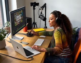 A woman in her home working at her desk with MacBook Pro connected to two external ekraans, wearing headphones and speaking into a microphone