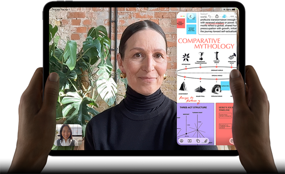 iPad Pro, front exterior, landscape orientation, space black color, rounded corners, black display bezel, hands holding left and right sides, display showing a FaceTime conversation being live translated, and an educational app showing a presentation of "Comparative Mythology" with text, diagrams, and handwritten notes